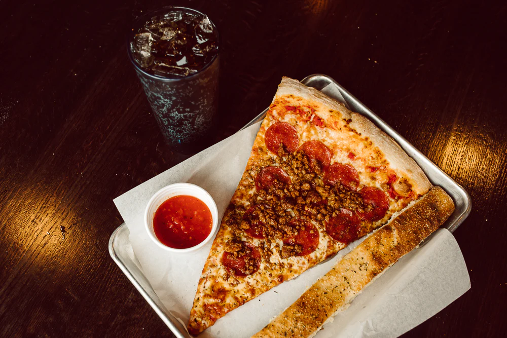 A photo of a slice of pizza, a breadstick and sauce, and a soft drink at Dough Co Drake.