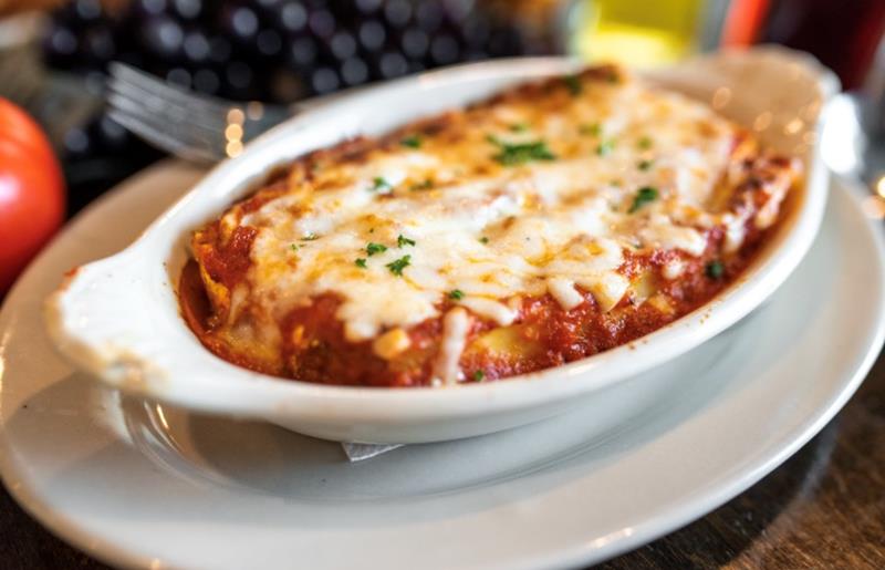 A pasta dish from Portofinos, featuring melted cheese on top. The dish is served in a white ceramic bowl with a decorative rim, garnished with green herbs. The pasta is shaped like shells and is coated in a red tomato sauce."