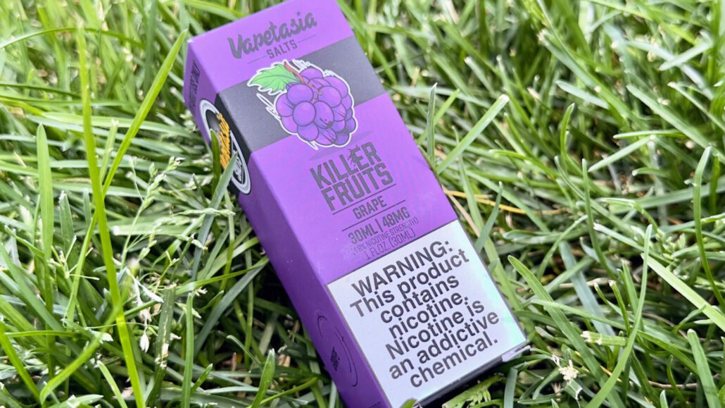 Featured image for the post, Vapetasia's Grape Nicotine Salt Review: These Killer Fruits are Tasty AF!" with the product box against a grass background.