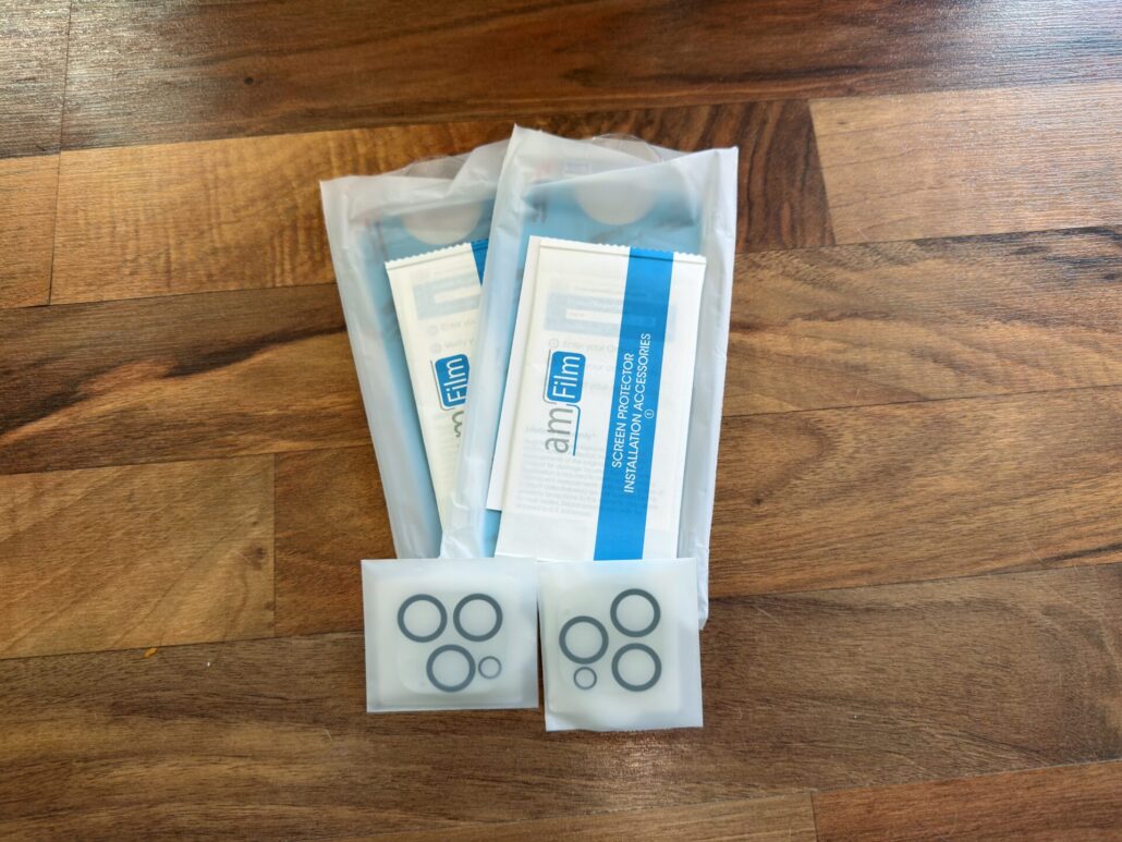 amFilm OneTouch iPhone 16 Pro Max kit contents laid out on a table, including two screen protector packs, cleaning wipes, and two sets of camera lens covers.