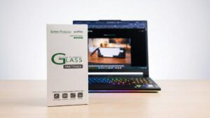 amFilm OneTouch Tempered Glass Screen Protector packaging standing upright on a desk next to a gaming laptop with an RGB keyboard.
