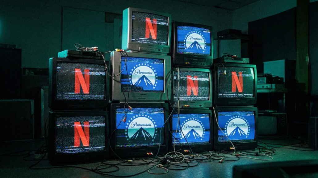 A stack of vintage CRT televisions in a dark room with entangled cables, featuring glitching and distorted screens showing alternating Netflix and Paramount logos. The image symbolizes the chaotic corporate battle and Paramount's hostile bid for Warner Bros. Discovery against Netflix.