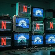 A stack of vintage CRT televisions in a dark room with entangled cables, featuring glitching and distorted screens showing alternating Netflix and Paramount logos. The image symbolizes the chaotic corporate battle and Paramount's hostile bid for Warner Bros. Discovery against Netflix.