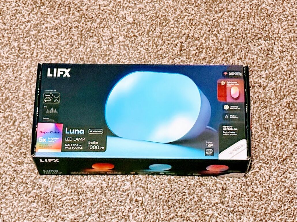 LIFX Luna retail box photographed from above on a carpet, with the lamp and its 1,000 lumen rating printed on the front panel